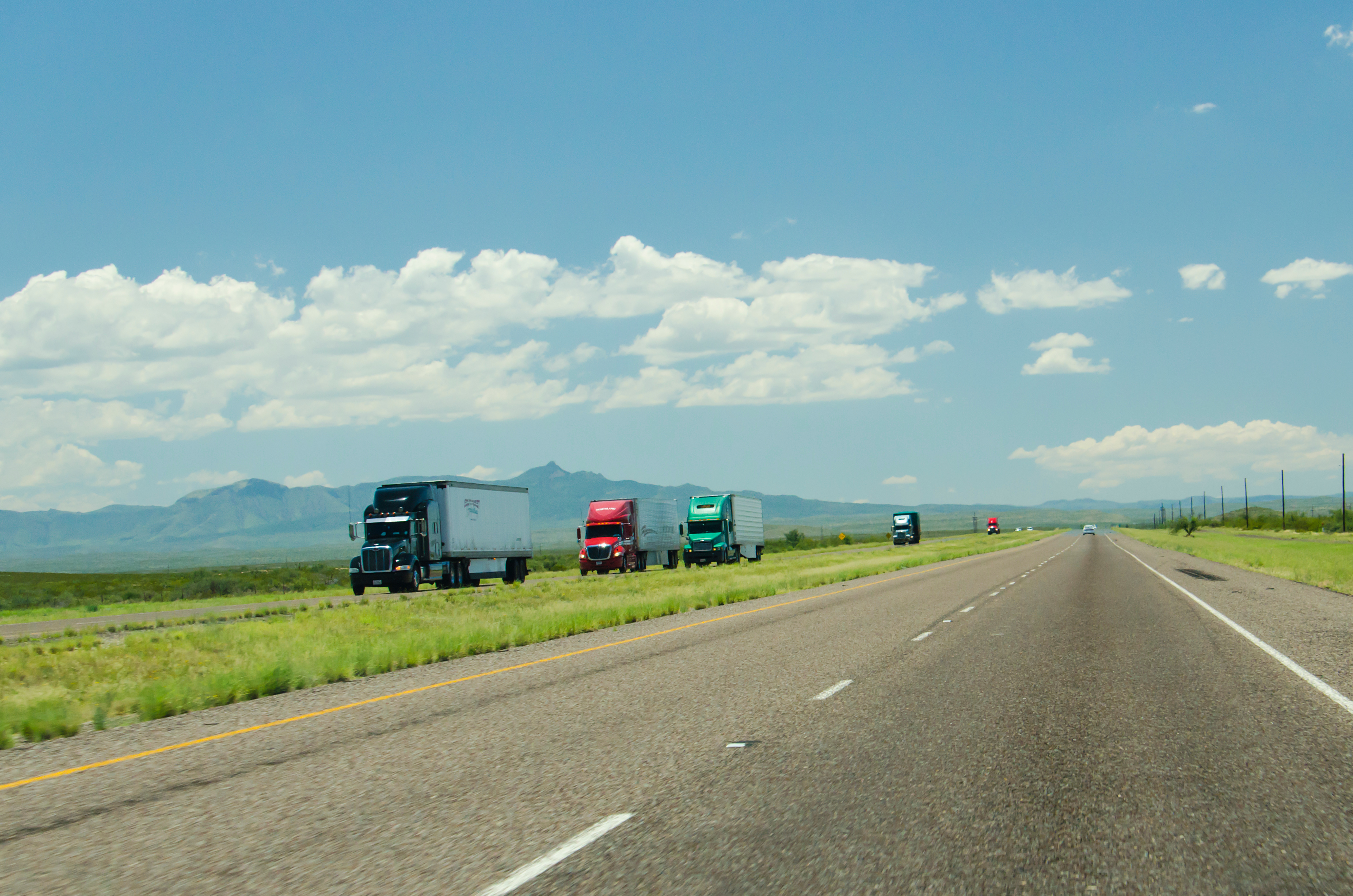 A row of semi-trucks travel down a wide, flat highway in western United States (Adobe Stock).