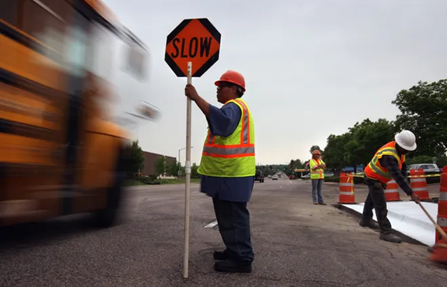 Several construction workers work on a road while another worker wearing a safety vest holds up a "Slow" sign to passing traffic.