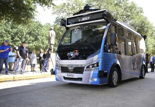 Press and onlookers gather on a crowded sidewalk in front of a silver and blue autonomous bus in Louisiana (U.S. DOT).