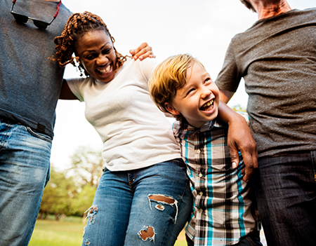 An intergenerational group of community members stands in a park smiling with their arms behind one another's backs.