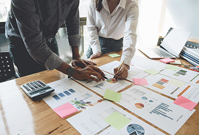 With a calculator at hand, two business people lean over a table and scrutinize multiple printed pages showing bar graphs and pie charts.