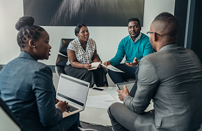 Four professional colleagues sit in a circle with their papers and laptops, debating an issue.