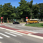 A street with a raised median and a marked crosswalk at a roundabout with yield signs and a school bus stopped for boarding passengers at the opposite entrance of the roundabout.