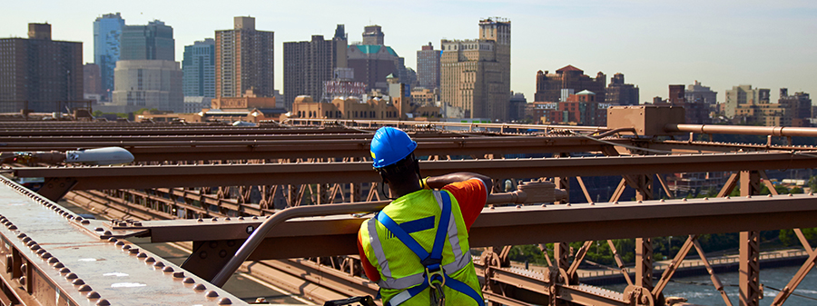 A man wearing a hard hat and safety harness works on bridge supports from the bucket of a cherry picker.