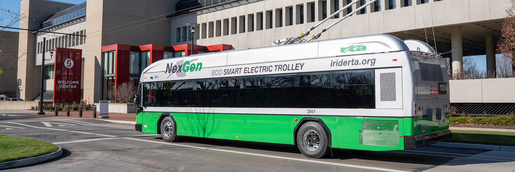 An electric trolleybus on a roadway in front of a building. The trolleybus is connected to overhead catenary wires.