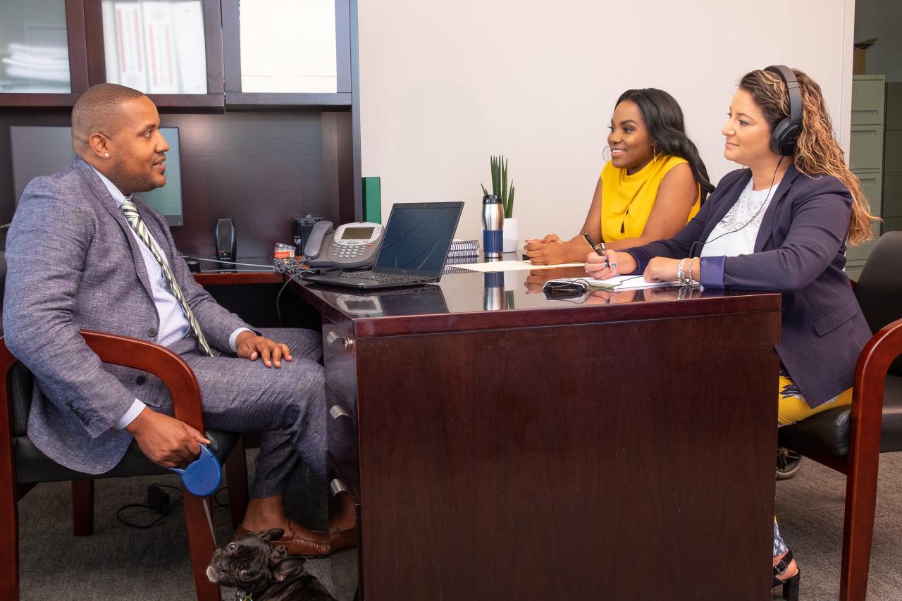 "Three individuals with disabilities of various backgrounds sit around an office desk. A small dog sits next to the desk."