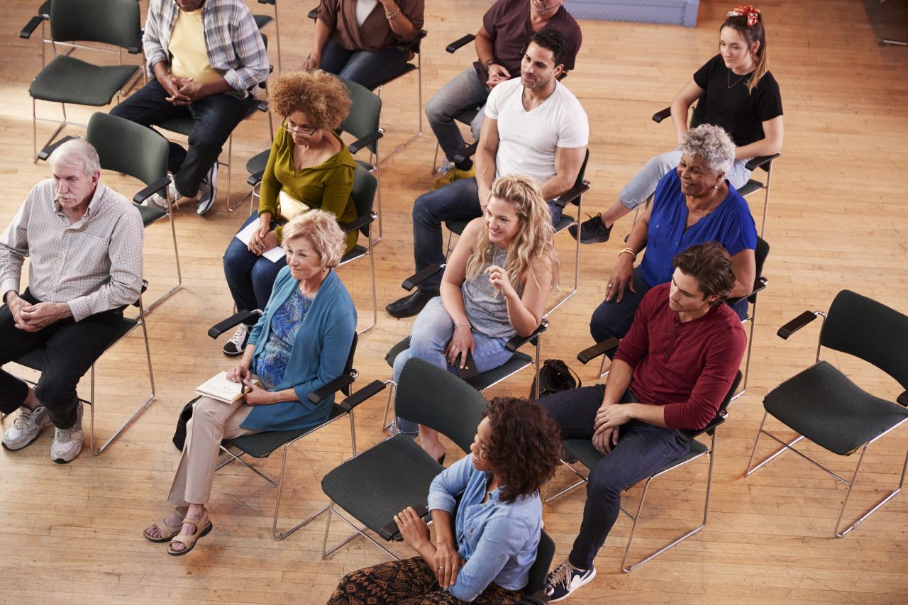 A group of people are seated in spaced-out chairs on a wood floor. The people and chairs are all facing the same direction. Some people are smiling, and some have notepads in their laps.