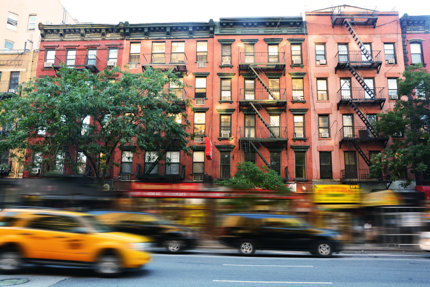 Cars and a taxi on a city street in front of a row of 5-story apartment buildings. 