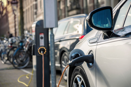 Close up of a charging cable plugged into an electric vehicle. The charger pedestal, a bike rack filled with bikes, and another parked vehicle are in the background.