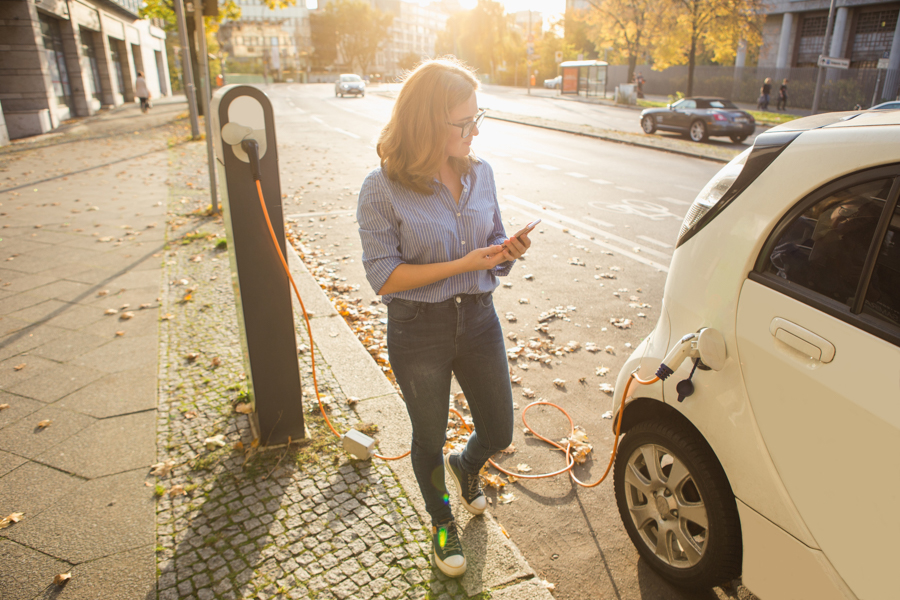 A woman stands on the edge of the sidewalk next to a parked electric vehicle that is plugged into a charger. 
