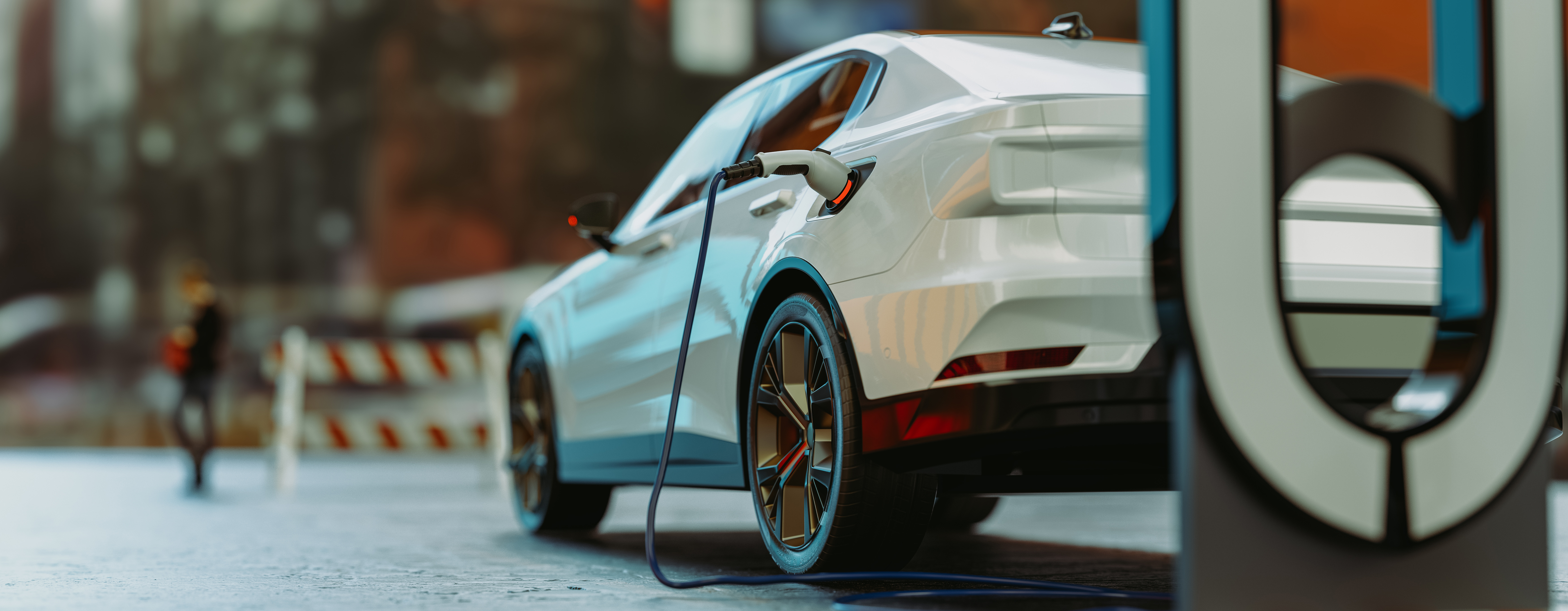 A parked car with a plugged-in charging cable. The pedestal charger is in the foreground. In the background a person walks next to a traffic barricade.