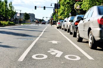 Roadway with bike lane running along parked cars on the side of the street and person crossing road in the distance.