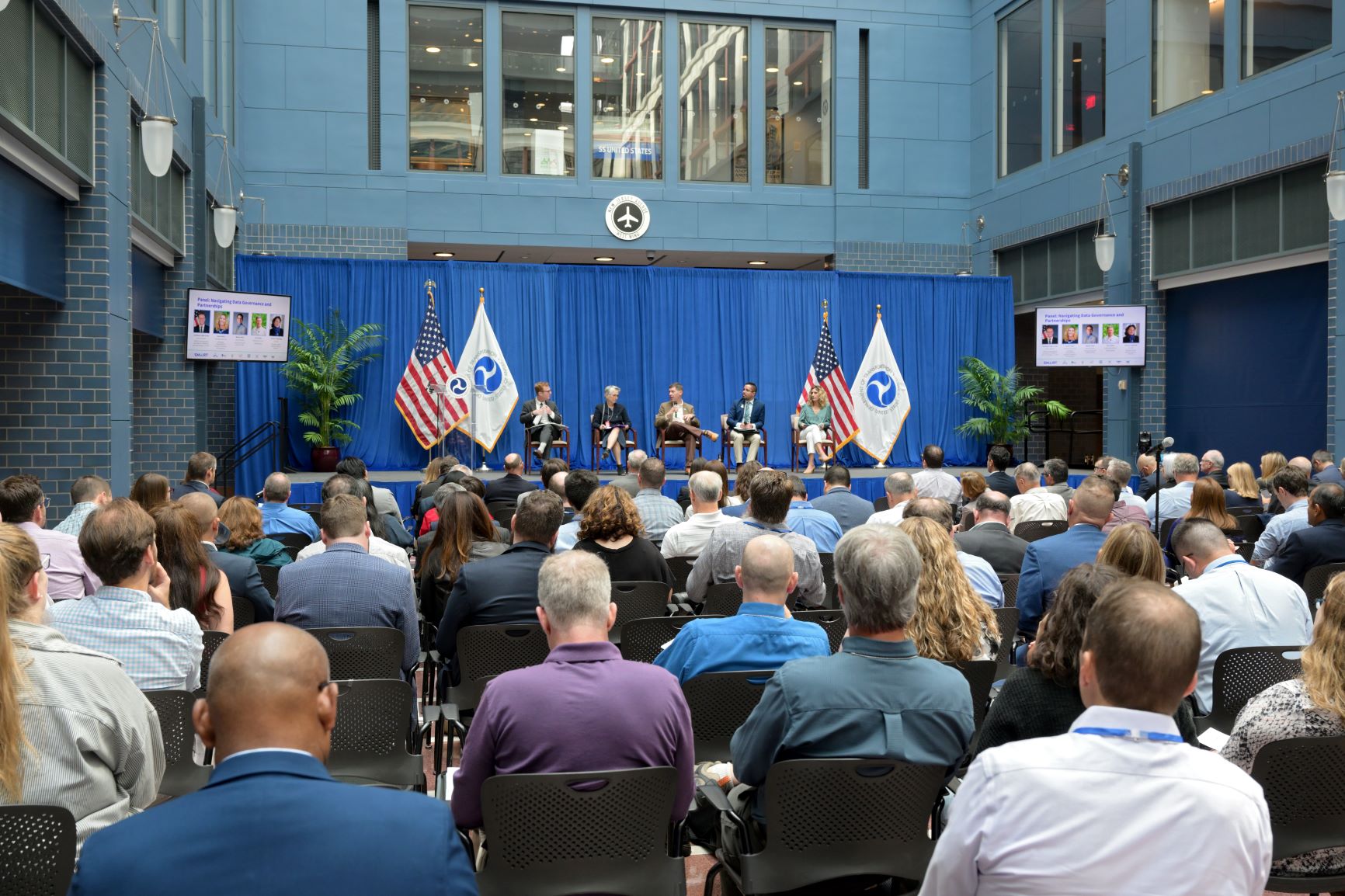 Five panelists sit at the front of an auditorium with many people watching from the audience.