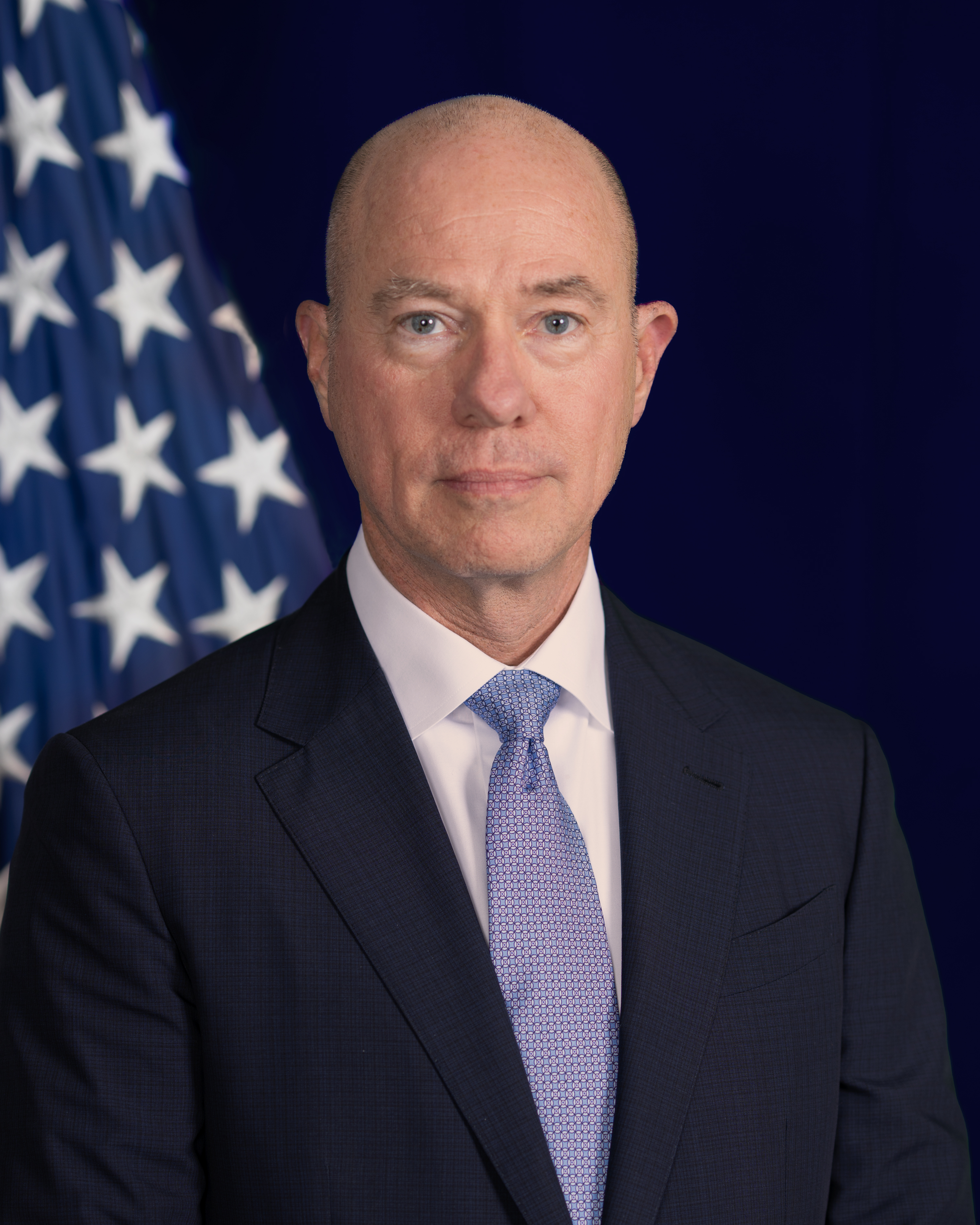 Man wearing a light blue tie standing in front of an American Flag.