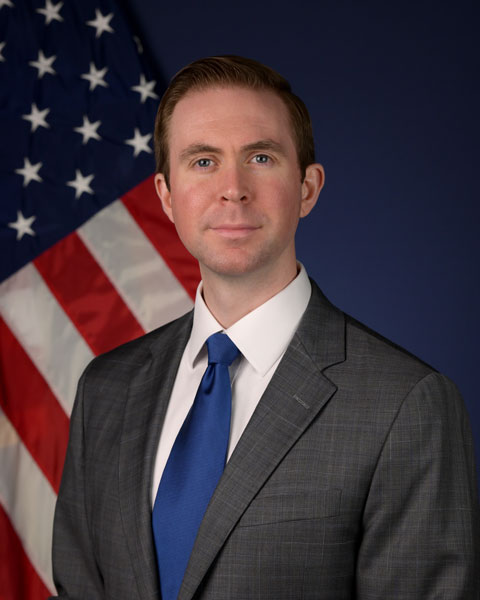 Middle aged man with brown hair and blue tie in front of American Flag.
