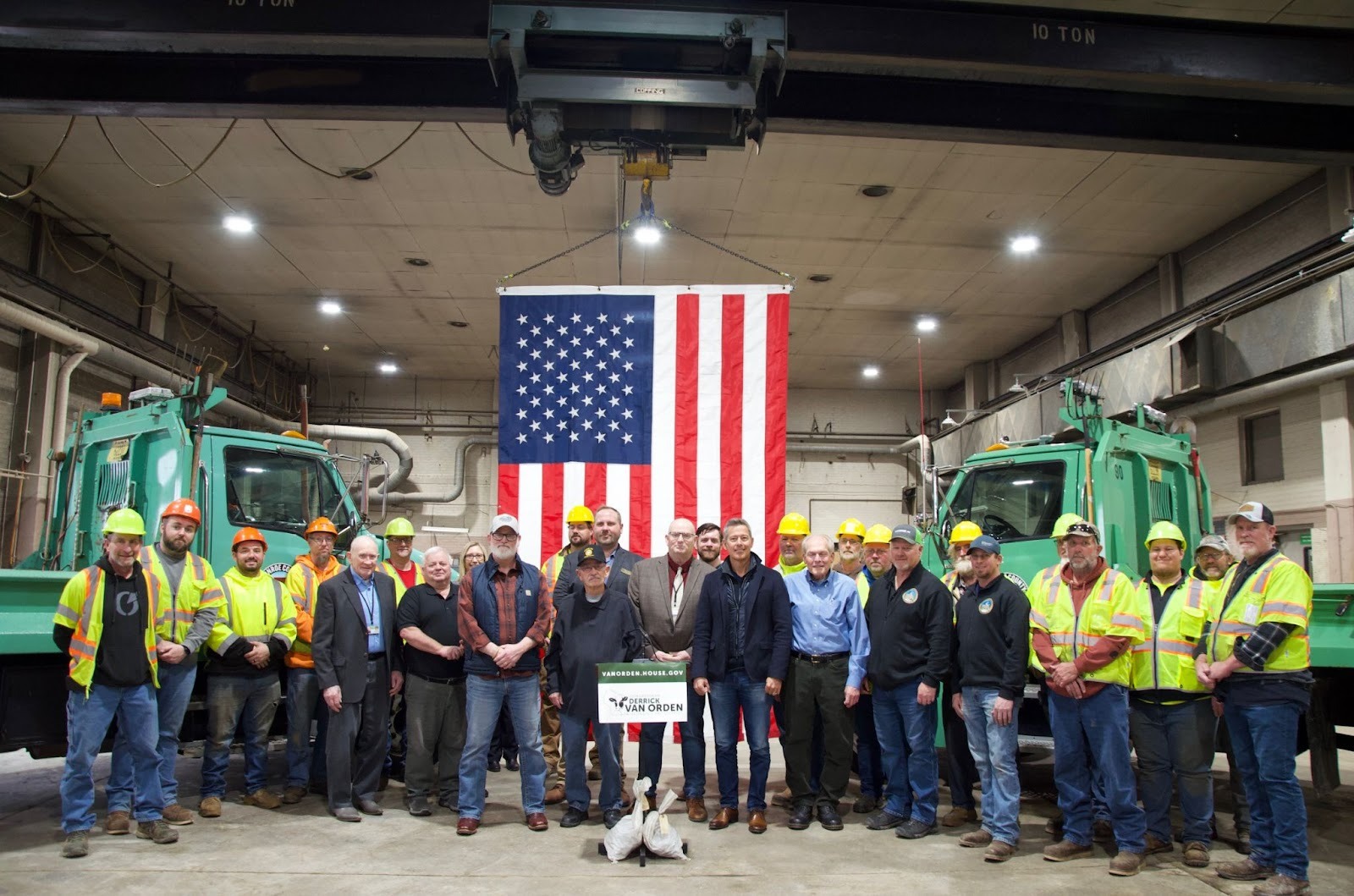 Secretary Duffy and Van Orden with workers in front of an American flag and trucks