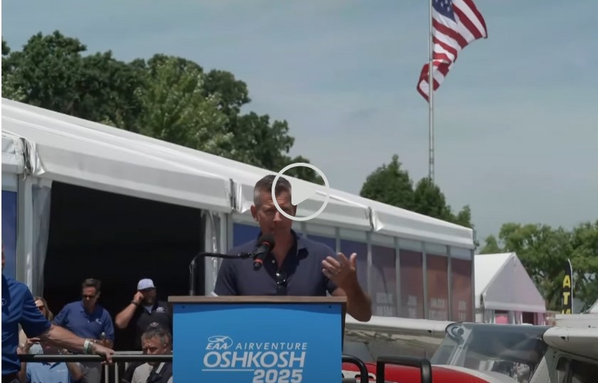 Secretary Duffy in front of Oshkosh 2025 podium with player icon at the center of the image