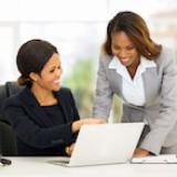 A woman looks at a laptop as another looks on.