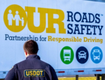 A USDOT safety inspector stands beside a semi trailer that reads: Our roads, our safety. Partnership for responsible driving.