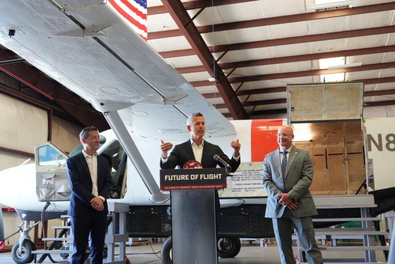Secretary Duffy speaking at podium with airplane behind him